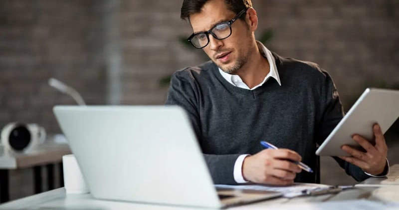 man with glasses working on a laptop and a notepad